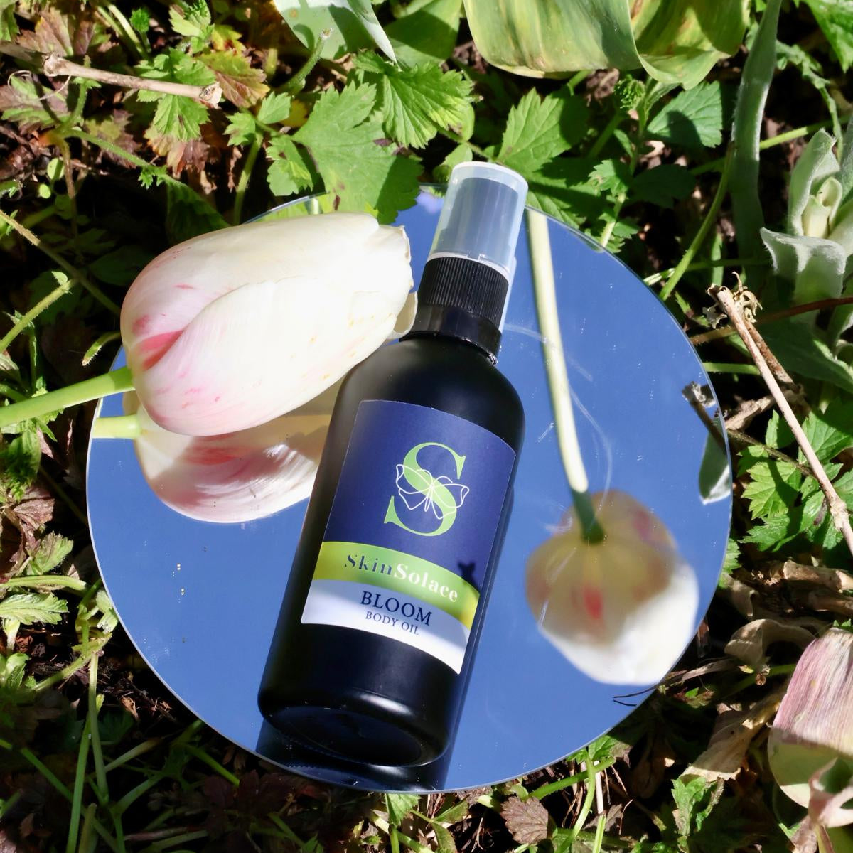 Skincare bottle labeled 'Bloom' on a blue mirror with flowers and greenery in the background